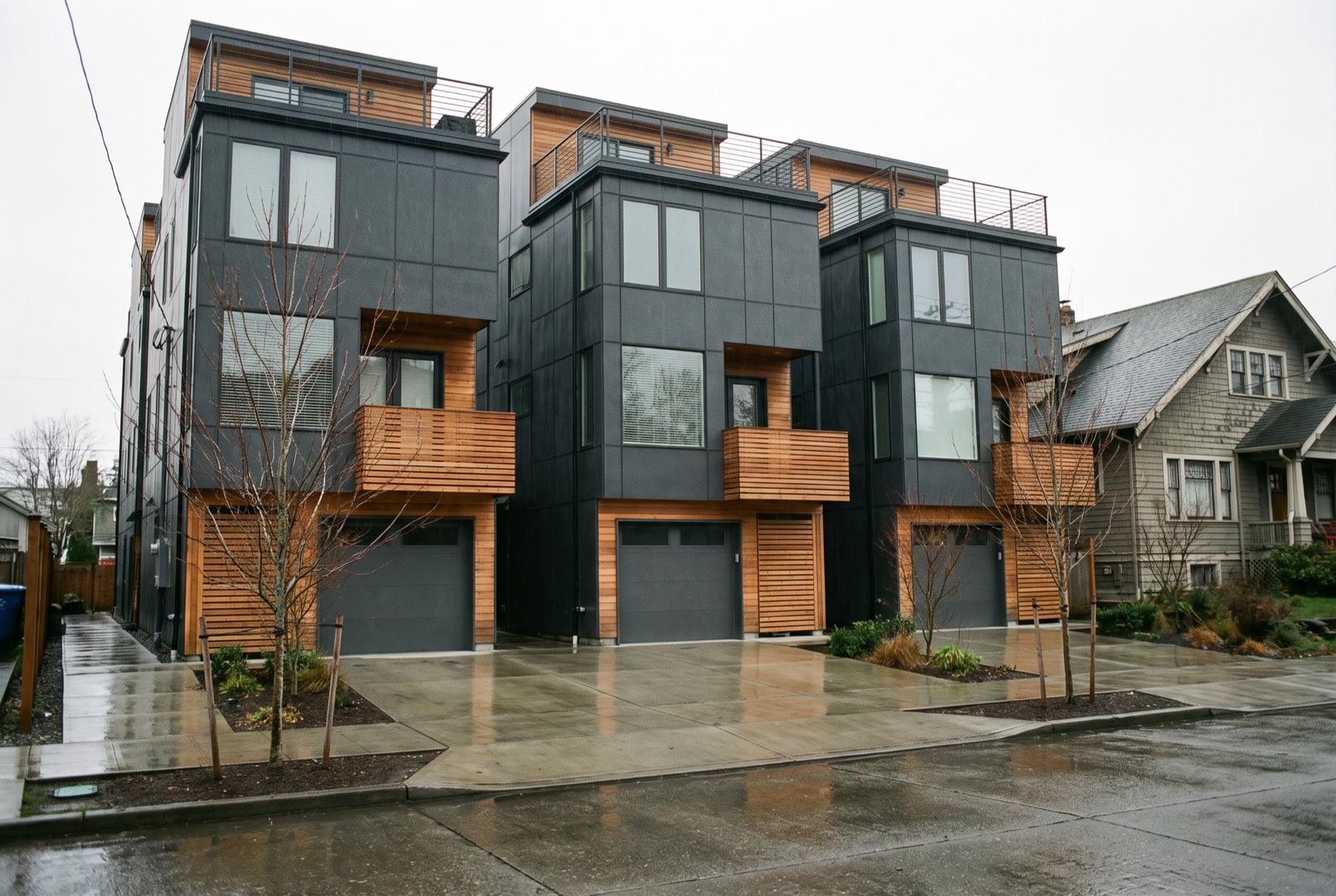 A row of new three-story townhomes with dark metal siding and cedar balconies above a shared concrete driveway on a rainy afternoon